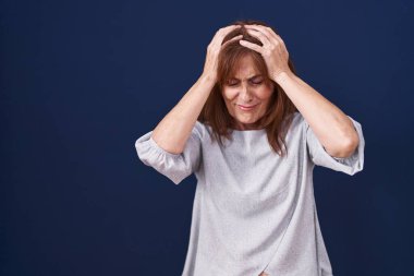Orta yaşlı bir kadın mavi arka planda dikiliyor. Baş ağrısı, çaresiz ve stresli. Ağrı ve migren yüzünden. Eller başının üstünde.. 