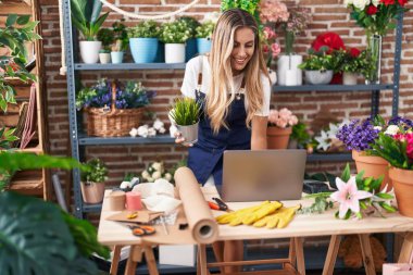 Young blonde woman florist using laptop holding plant at florist