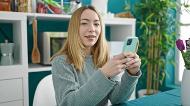 Genç sarışın kadın yemek odasındaki masada oturan akıllı telefonu kullanıyor.