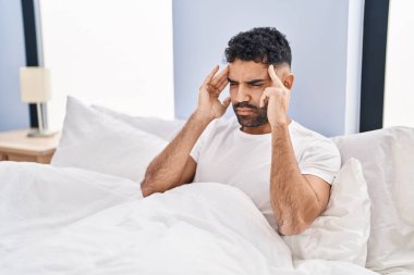 Young hispanic man suffering for headache sitting on bed at bedroom