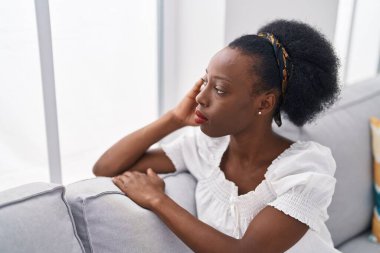 Kanepede oturan Afro-Amerikalı kadın ciddi bir ifadeyle evde oturuyor.