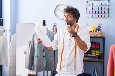 Genç İspanyol terzi akıllı telefondan konuşuyor giyim fabrikasında kıyafet tasarımı yapıyor.