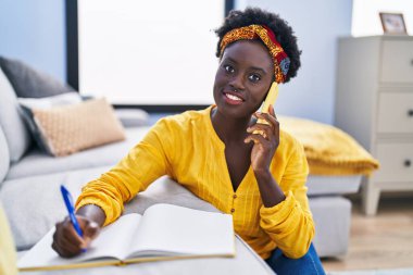 Young african american woman writing on notebook talking on the smartphone at home
