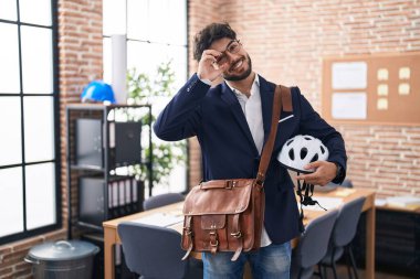 Ofiste sakallı, bisiklet kasklı, gülümseyen, mutlu bir İspanyol adam. Elleri gözlerinde, parmaklarının arasından bakıyor. 