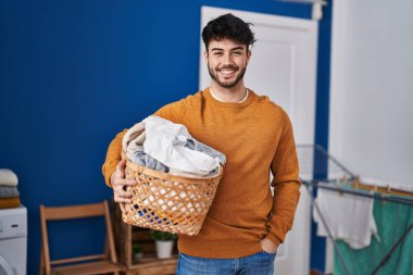 Çamaşır odasında sakallı İspanyol bir adam çamaşır sepetini tutuyor. Olumlu ve mutlu görünüyor. Kendine güvenen bir gülümsemeyle dişlerini gösteriyor. 