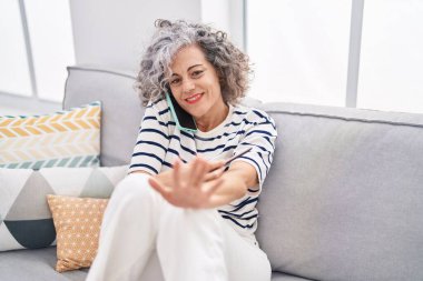 Orta yaşlı, kır saçlı, akıllı telefondan konuşan ve evde manikür yapan bir kadın.