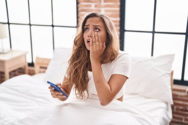 Genç, güzel, İspanyol bir kadın yatak odasında endişeli bir ifadeyle akıllı telefon kullanıyor.