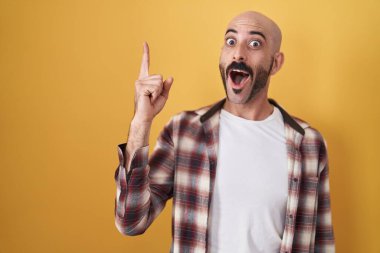 Hispanic man with beard standing over yellow background pointing finger up with successful idea. exited and happy. number one. 