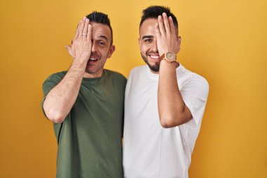 Sarı arka planda duran eşcinsel çift bir gözünü eliyle kapatıyor, yüzünde kendine güvenen bir gülümseme ve sürpriz bir duygu var.. 
