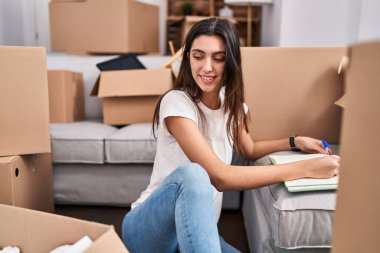 Young beautiful hispanic woman writing on notebook sitting on floor at new home