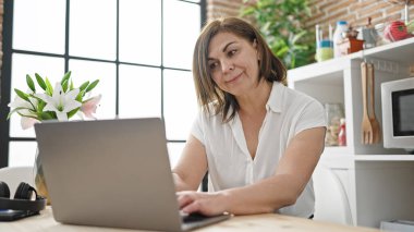 Orta yaşlı İspanyol kadın yemek odasında dizüstü bilgisayar kullanıyor.