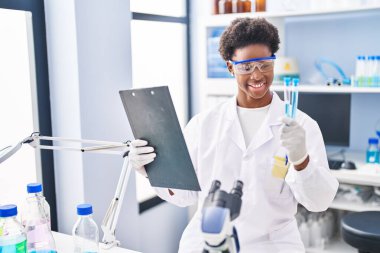 African american woman wearing scientist uniform reading document holding test tubes at laboratory