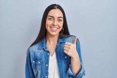 Mavi arka planda dikilen İspanyol kadın elleriyle mutlu bir jest yapıyor. Kameraya bakıp başarıyı gösteren ifade. 