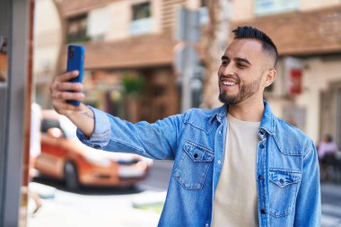 Young hispanic man smiling confident making selfie by the smartphone at street