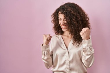 Hispanic woman with curly hair standing over pink background very happy and excited doing winner gesture with arms raised, smiling and screaming for success. celebration concept. 