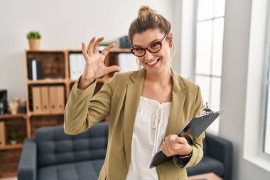 Muayenehanede çalışan genç bir kadın gülümsüyor ve el hareketleriyle hareket ediyor. Küçük boy işareti yapıyor, parmaklarına bakıyor ve kameraya bakıyor. ölçüm kavramı. 