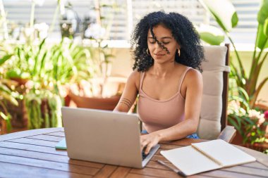 Young hispanic woman using laptop sitting on table at home terrace
