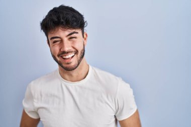 Hispanic man with beard standing over white background winking looking at the camera with sexy expression, cheerful and happy face. 