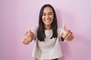 Young hispanic woman standing over pink background approving doing positive gesture with hand, thumbs up smiling and happy for success. winner gesture. 