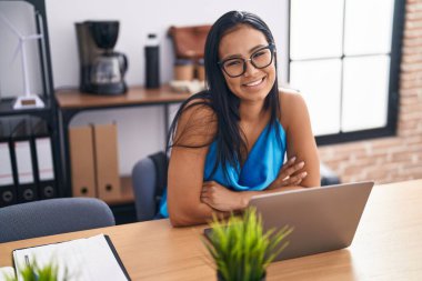 Young beautiful latin woman business worker using laptop sitting with arms crossed gesture at office
