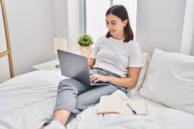Young hispanic woman sitting on bed using laptop studying at bedroom