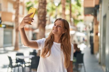 Genç sarışın kız, kendinden emin gülümsüyor. Sokakta akıllı telefonun yanında özçekim yapıyor.