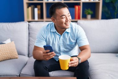 Genç Çinli adam akıllı telefon kullanıyor. Evde kahve içiyor.