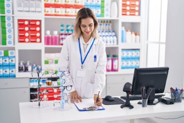 Young woman pharmacist smiling confident writing on document at pharmacy