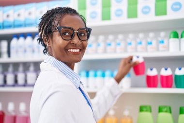 Middle age african american woman pharmacist smiling confident holding toothpaste on shelving at pharmacy
