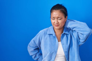 Asyalı genç bir kadın mavi arka planda duruyor. Boyun ağrısı çekiyor, eliyle boynuna dokunuyor, kas ağrısı çekiyor. 