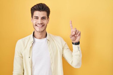 Sarı arka planda duran İspanyol genç adam bir numarayı gösterip işaret ederken kendinden emin ve mutlu bir şekilde gülümsüyor.. 