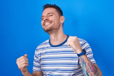 Young hispanic man standing over blue background dancing happy and cheerful, smiling moving casual and confident listening to music 