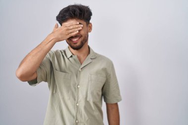 Beyaz arka planda duran sakallı Arap bir adam. Gülümsüyor ve gülümsüyor. Gözlerini şaşkınlıkla kapatıyor. kör kavram. 