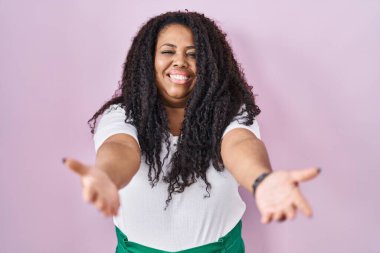 Artı, pembe arka planda duran boyda bir İspanyol kadın kameraya bakıp sarılmak için kollarını açmış gülümsüyor. Mutluluğu kucaklayan neşeli bir ifade.. 