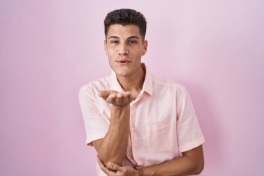 Young hispanic man standing over pink background looking at the camera blowing a kiss with hand on air being lovely and sexy. love expression. 