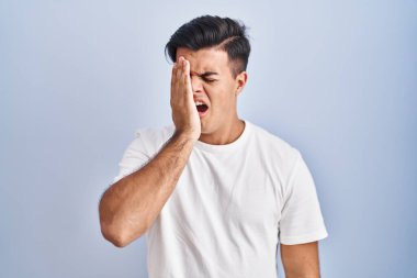 Hispanic man standing over blue background yawning tired covering half face, eye and mouth with hand. face hurts in pain. 