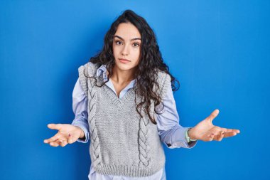 Mavi arka planda duran esmer bir kadın. Hiçbir fikri olmayan, kolları açık, kafası karışık bir kadın.. 