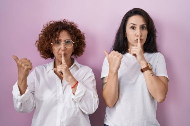 İspanyol anne ve kızı pembe arka plan üzerine günlük beyaz t-shirt giyip parmakları dudaklarını yana doğru gösteren parmaklarıyla sessiz olmak istiyorlar. sessizlik ve gizli konsept. 