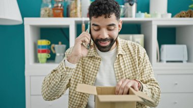 Genç İspanyol adam, yemek odasındaki karton kutuları açarak akıllı telefondan konuşuyor.