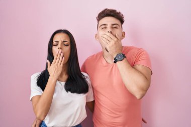 Young hispanic couple standing over pink background bored yawning tired covering mouth with hand. restless and sleepiness. 