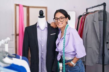 Young beautiful hispanic woman tailor smiling confident leaning on manikin at tailor shop