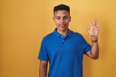 Sarı arka planda duran İspanyol genç adam beş numarayı gösterip işaret ederken kendinden emin ve mutlu gülümsüyor.. 