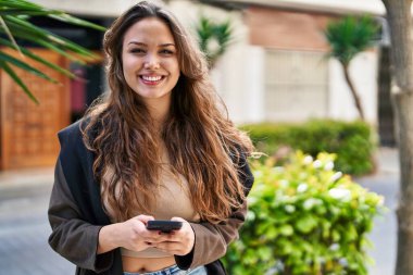Genç, güzel, İspanyol bir kadın sokakta akıllı telefon kullanarak gülümsüyor.