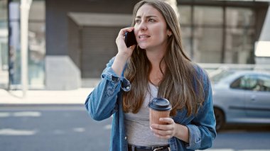 Young beautiful hispanic woman talking on smartphone drinking coffee at street