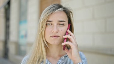 Young blonde woman talking on smartphone with serious expression at street