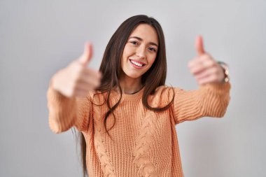 Genç esmer kadın beyaz arka planda durup olumlu jestler yapmayı onaylıyor, gülümsüyor ve başarı için mutlu. kazanan jest. 