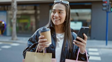 Genç ve güzel İspanyol kadın akıllı telefon kullanıyor. Elinde alışveriş poşetleri ve sokakta kahve tutuyor.