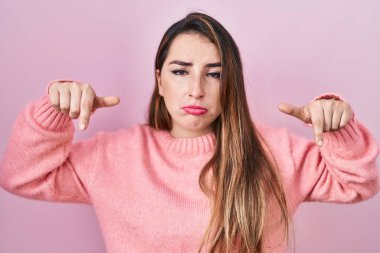 Young hispanic woman standing over pink background pointing down looking sad and upset, indicating direction with fingers, unhappy and depressed. 