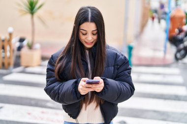 Genç, güzel, İspanyol bir kadın sokakta akıllı telefon kullanarak gülümsüyor.