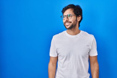 Mavi arka planda duran yakışıklı latin adam yüzünde gülümseme ve doğal ifadeyle yan yana bakıyor. Kendinden emin gülen. 